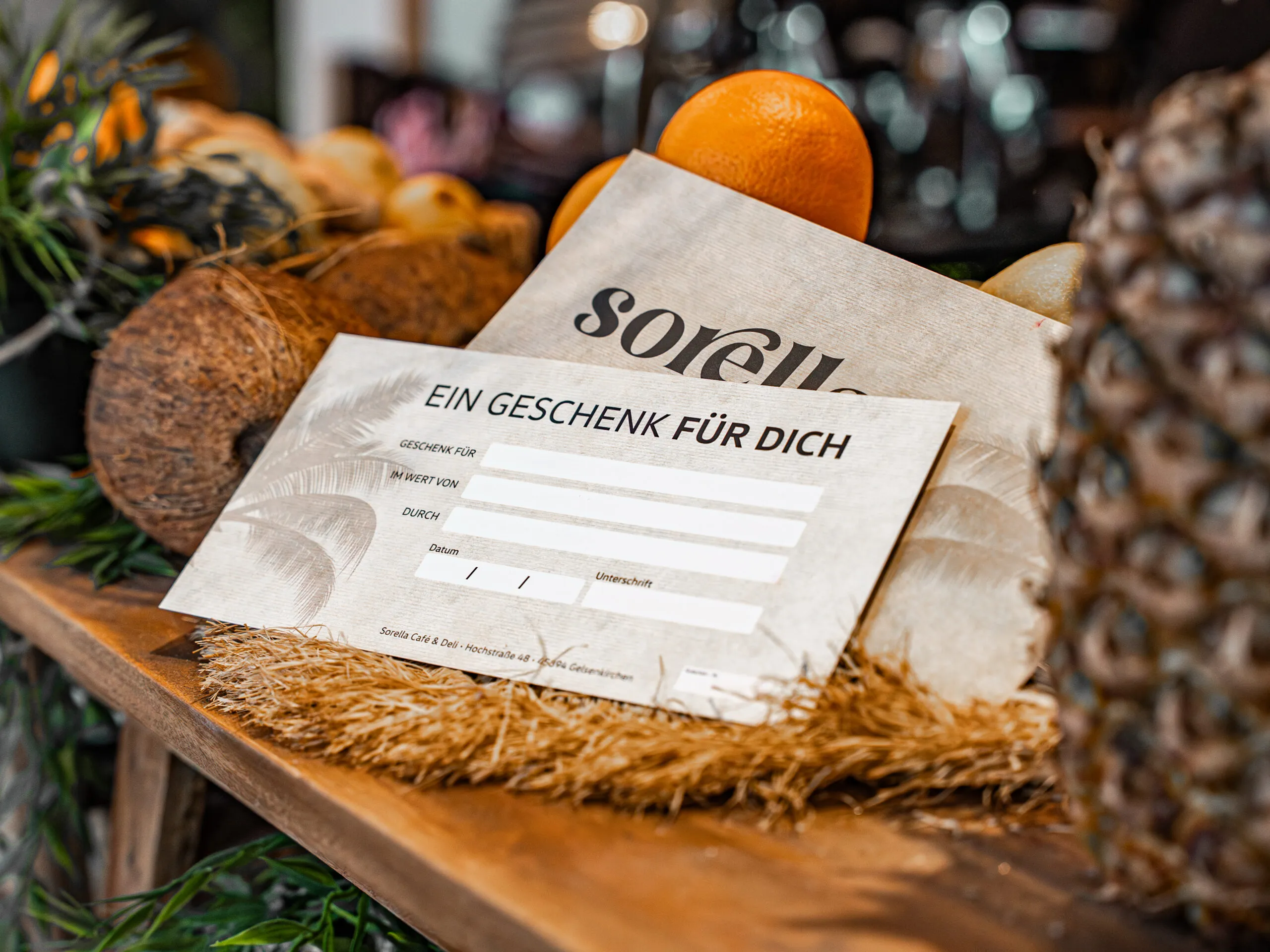 Sorella Gutschein auf einer Holzbank neben Ananas, Kokosnuss und Orangen