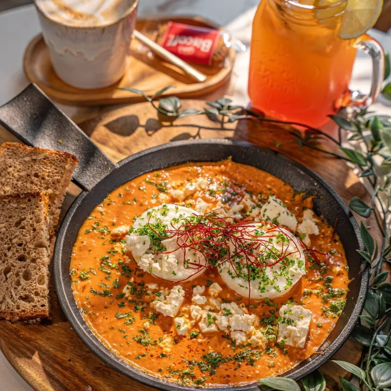 Shakshuka mit Feta und Kräutern in der Pfanne, daneben Kaffee und Eistee